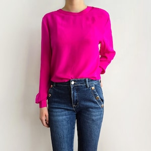 Könnte beinhalten: Eine leuchtend pinkfarbene Langarmbluse kombiniert mit dunkelblauen Jeans. Die Bluse hat einen runden Ausschnitt und die Jeans sind mit goldfarbenen Knöpfen versehen. Ein einfaches und stilvolles Outfit.