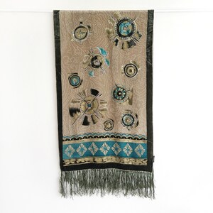 May include: Tan scarf with a repeating pattern of circular designs in black, gold, and teal. The scarf has a black border and a teal and gold patterned band near the bottom. The scarf is finished with a fringe.