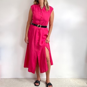 May include: A sleeveless, hot pink midi dress with a button-down front and a high slit. The dress is cinched at the waist with a black belt featuring a gold buckle. The model is wearing black sandals.