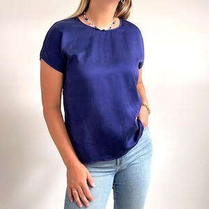 May include: A navy blue, short-sleeved blouse with a round neckline. The top is made of a smooth, shiny fabric. The model is wearing light blue jeans and a necklace.