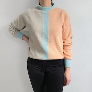 May include: A color-blocked sweater featuring a light beige and peach color scheme, bisected by a vertical light blue stripe. The sweater has a mock neck and light blue cuffs and trim.