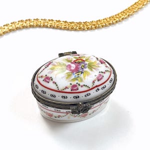 Puede incluir: Caja de baratijas ovalada de porcelana blanca con diseño floral. La tapa presenta flores rosas, moradas y amarillas con hojas verdes. La caja tiene un borde rojo con pequeños números negros y un cierre metálico.