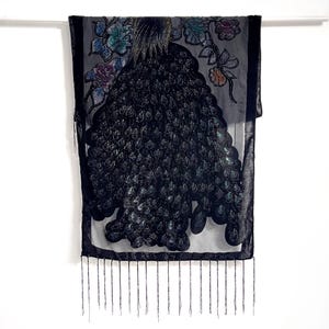 Puede incluir: Una bufanda negra con un diseño de pavo real. La bufanda presenta un diseño detallado de pavo real con acentos iridiscentes y adornos florales en tonos de azul, morado y dorado. El borde inferior tiene flecos.
