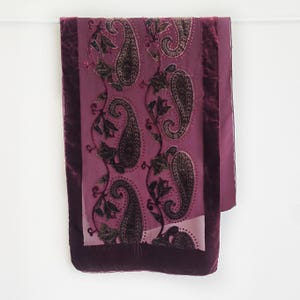 Puede incluir: Bufanda decorativa con estampado de cachemir en tonos morados y burdeos. La bufanda presenta un panel central con grandes diseños de cachemir y un borde de terciopelo burdeos oscuro. Los elementos de cachemir son de un tono más oscuro.