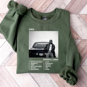 May include: A green sweatshirt with a black and white image of a man standing next to a car. The image is a record cover for Kendrick Lamar's album "GNX". The album title is printed in white on the sweatshirt. The album tracklist is printed below the image.