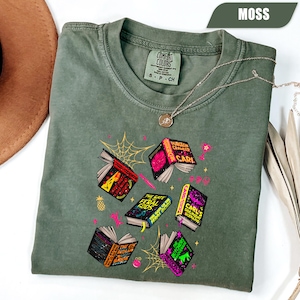 May include: Moss green Comfort Colors t-shirt with a graphic of open books, a spiderweb, and other decorative elements. The books have titles like "Dungeon Crawler Carl" and "The Gate of the Feral Gods". A silver necklace is also visible.