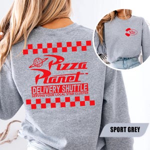 May include: A sport grey crewneck sweatshirt featuring a red Pizza Planet logo on the back with checkered borders and the text Delivery Shuttle Serving Your Local Star Cluster. The front chest displays a smaller red Pizza Planet logo graphic.