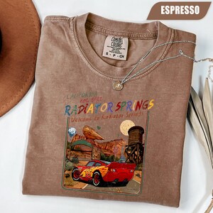 May include: An espresso-colored t-shirt featuring a graphic print of a red race car with the number 95, a desert landscape, and the text California Est. 2012 Radiator Springs Welcome to Radiator Springs. The shirt has a Comfort Colors brand label.