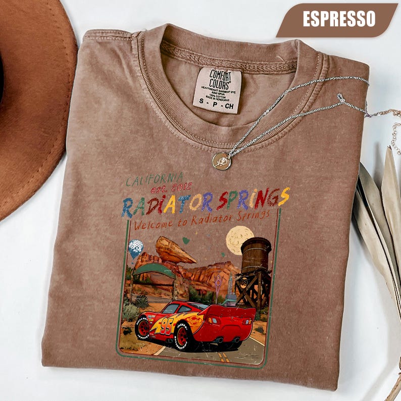 May include: An espresso-colored t-shirt featuring a graphic print of a red race car with the number 95, a desert landscape, and the text California Est. 2012 Radiator Springs Welcome to Radiator Springs. The shirt has a Comfort Colors brand label.