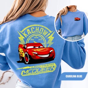 May include: A Carolina blue sweatshirt featuring a large graphic on the back of the red race car Lightning McQueen with the text KACHOW! and LIGHTNING MCQUEEN in yellow. A smaller version of the red race car is printed on the front left chest.