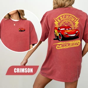 May include: A crimson t-shirt featuring a small graphic of the red race car Lightning McQueen on the front left chest. The back of the shirt displays a large yellow graphic of Lightning McQueen with the text KACHOW and LIGHTNING MCQUEEN.