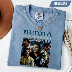 Puede incluir: Una camiseta Comfort Colors azul claro con un gráfico de Pedro Pascal y el texto "PEDRO Pascal". La camiseta tiene cuello redondo y mangas cortas. Un collar dorado con un colgante está drapeado sobre la camiseta.