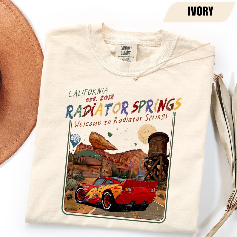 May include: An ivory t-shirt featuring a graphic print of a red race car with the number 95. The text reads California est. 2012, Radiator Springs, and Welcome to Radiator Springs. The design includes a desert landscape, a water tower, and a rock formation.