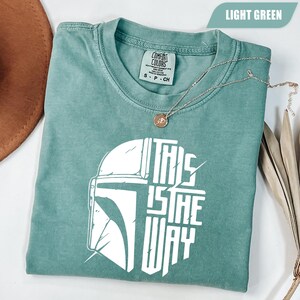 May include: Light green t-shirt with the text "THIS IS THE WAY" in white, along with a graphic of a helmet. A gold necklace with a pendant is draped over the shirt. The shirt is folded and has a tag that says "Comfort Colors".