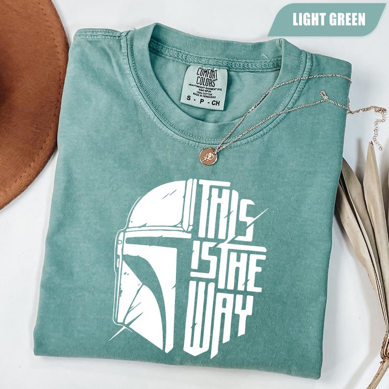 May include: Light green t-shirt with the text "THIS IS THE WAY" in white, along with a graphic of a helmet. A gold necklace with a pendant is draped over the shirt. The shirt is folded and has a tag that says "Comfort Colors".