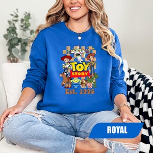 Puede incluir: Sudadera azul real con un gr&aacute;fico colorido de los personajes de Toy Story. El dise&ntilde;o incluye a Woody, Buzz Lightyear y otros personajes de la pel&iacute;cula, con el texto "Toy Story" y "Est. 1995".