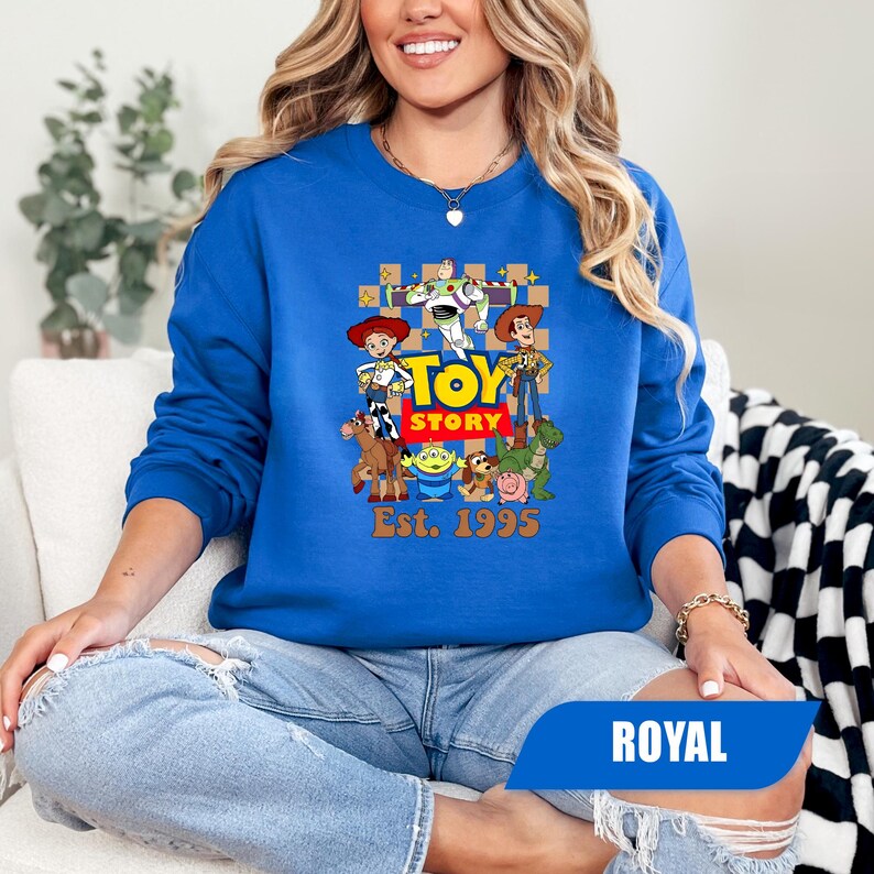 Puede incluir: Sudadera azul real con un gr&aacute;fico colorido de los personajes de Toy Story. El dise&ntilde;o incluye a Woody, Buzz Lightyear y otros personajes de la pel&iacute;cula, con el texto "Toy Story" y "Est. 1995".