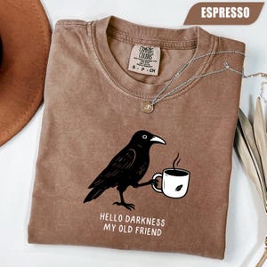 Peut inclure: T-shirt couleur expresso avec un dessin d'un corbeau noir tenant une tasse de café. L'inscription "Hello Darkness My Old Friend" est imprimée en blanc. Un collier argenté avec un pendentif est posé sur le t-shirt.