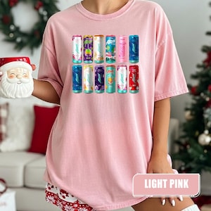 May include: A light pink t-shirt featuring a graphic print of twelve colorful Alani Nu energy drink cans arranged in two rows. The cans display various vibrant colors and fruit illustrations, with the brand name Alani clearly visible on each.