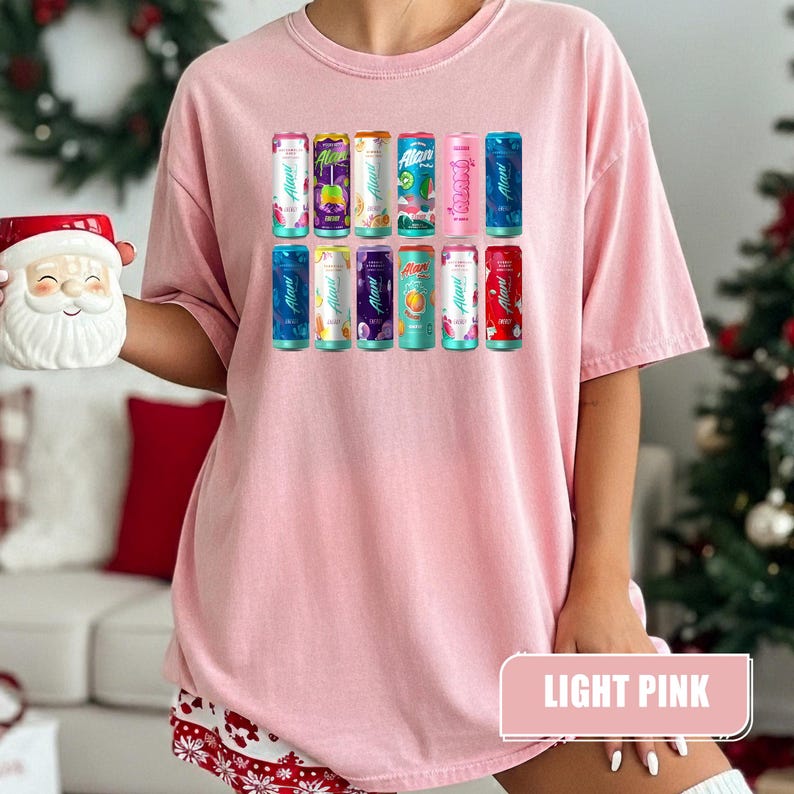 May include: A light pink t-shirt featuring a graphic print of twelve colorful Alani Nu energy drink cans arranged in two rows. The cans display various vibrant colors and fruit illustrations, with the brand name Alani clearly visible on each.