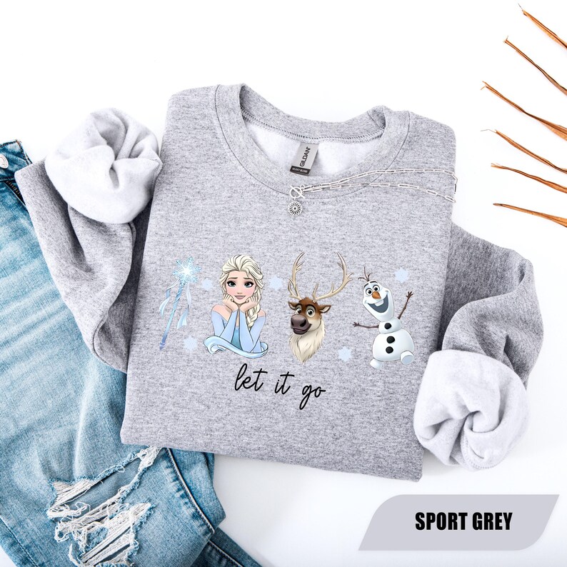 May include: Sport grey sweatshirt featuring Elsa, Sven, and Olaf from Frozen. The design includes the phrase "let it go" and a snowflake wand. The sweatshirt has a crew neck and long sleeves with white cuffs. The words "Sport Grey" are printed at the bottom.