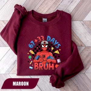 May include: Maroon sweatshirt featuring a Spider-Man graphic with the text "67+33 DAYS BRUH". The graphic includes a checkered background and various school-themed illustrations. The sweatshirt is a solid maroon color.