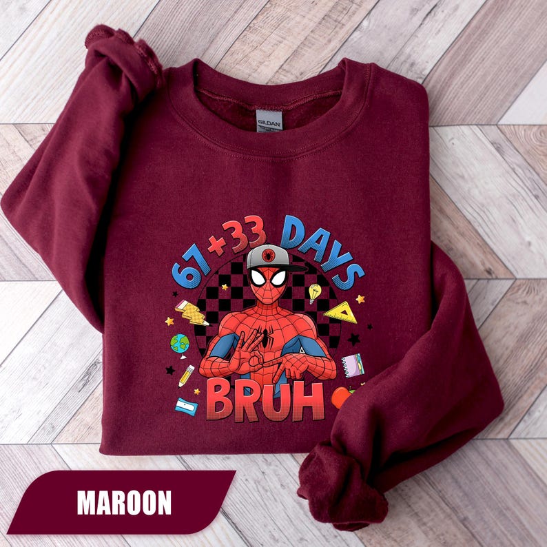 May include: Maroon sweatshirt featuring a Spider-Man graphic with the text "67+33 DAYS BRUH". The graphic includes a checkered background and various school-themed illustrations. The sweatshirt is a solid maroon color.