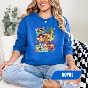 Puede incluir: Sudadera azul real con un gr&aacute;fico colorido de personajes de Toy Story. El dise&ntilde;o incluye im&aacute;genes de Woody, Buzz Lightyear y otros personajes. La palabra "ROYAL" est&aacute; impresa en una pancarta azul en la parte inferior.