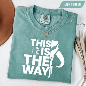 May include: A light green short-sleeve t-shirt featuring the white text "THIS IS THE WAY" alongside a graphic of a helmet and a stylized silhouette. The shirt has a crew neckline and a Comfort Colors brand label printed on the inside collar area.