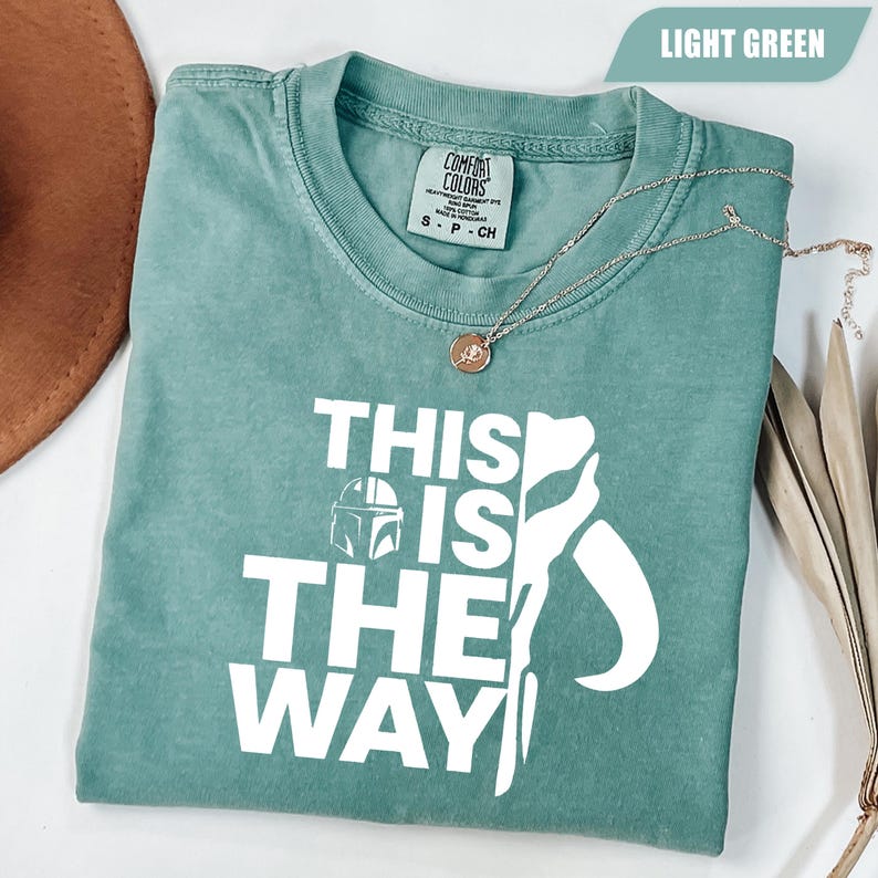 May include: A light green short-sleeve t-shirt featuring the white text "THIS IS THE WAY" alongside a graphic of a helmet and a stylized silhouette. The shirt has a crew neckline and a Comfort Colors brand label printed on the inside collar area.