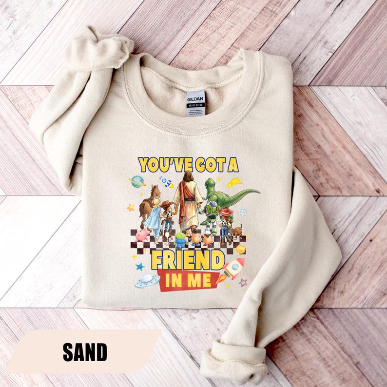 May include: A sand-colored crewneck sweatshirt featuring a graphic print of a central figure surrounded by various animated characters standing on a checkered floor. The text "YOU'VE GOT A FRIEND IN ME" is printed in yellow and red above and below the characters.