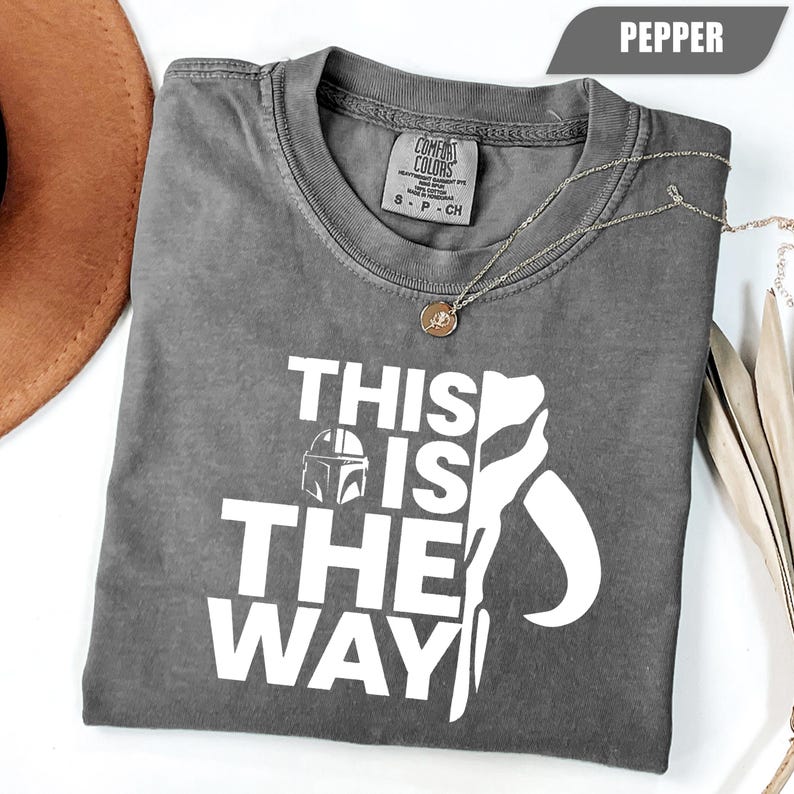 May include: A pepper-colored short-sleeve t-shirt featuring the white text "THIS IS THE WAY" and a graphic of a Mandalorian helmet. The shirt has a Comfort Colors brand label at the inner neckline and a thin gold-toned chain necklace resting on top.