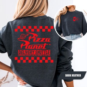 May include: A dark heather gray crewneck sweatshirt featuring a red graphic on the back with a checkered border, the text Pizza Planet, and the words DELIVERY SHUTTLE SERVING YOUR LOCAL STAR CLUSTER. A smaller red logo is on the front chest.