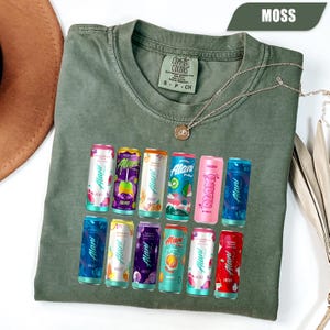 May include: A moss green crew neck t-shirt with a Comfort Colors label at the inner collar. The front features a graphic print of twelve colorful Alani energy drink cans arranged in two rows, showcasing various fruit-themed designs and labels.