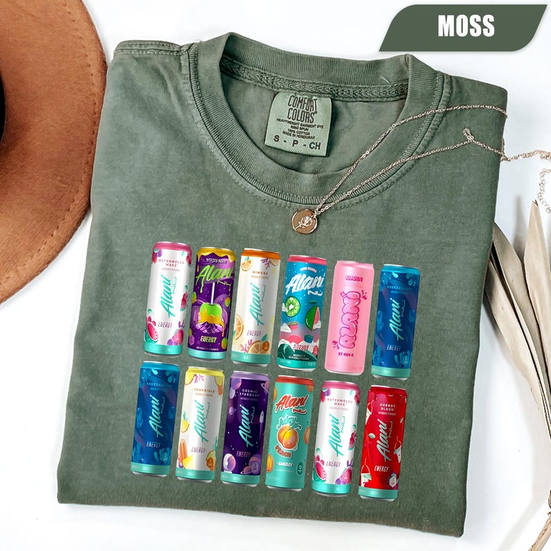 May include: A moss green crew neck t-shirt with a Comfort Colors label at the inner collar. The front features a graphic print of twelve colorful Alani energy drink cans arranged in two rows, showcasing various fruit-themed designs and labels.