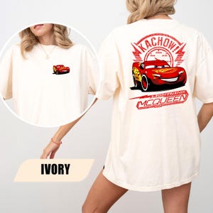 May include: An ivory t-shirt featuring a small graphic of a red race car on the front left chest. The back displays a large red graphic of the same race car with the text KACHOW! and LIGHTNING MCQUEEN printed in a distressed red font style.