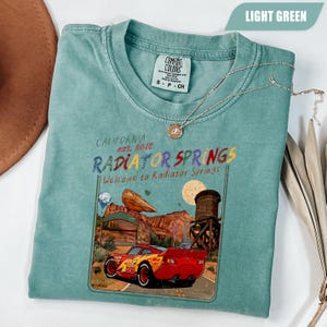 May include: A light green short-sleeve t-shirt featuring a graphic print of a red race car with the number 95, a desert landscape, a water tower, and the text California est. 2012 Radiator Springs Welcome to Radiator Springs on the front center.
