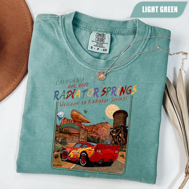 May include: A light green short-sleeve t-shirt featuring a graphic print of a red race car with the number 95, a desert landscape, a water tower, and the text California est. 2012 Radiator Springs Welcome to Radiator Springs on the front center.