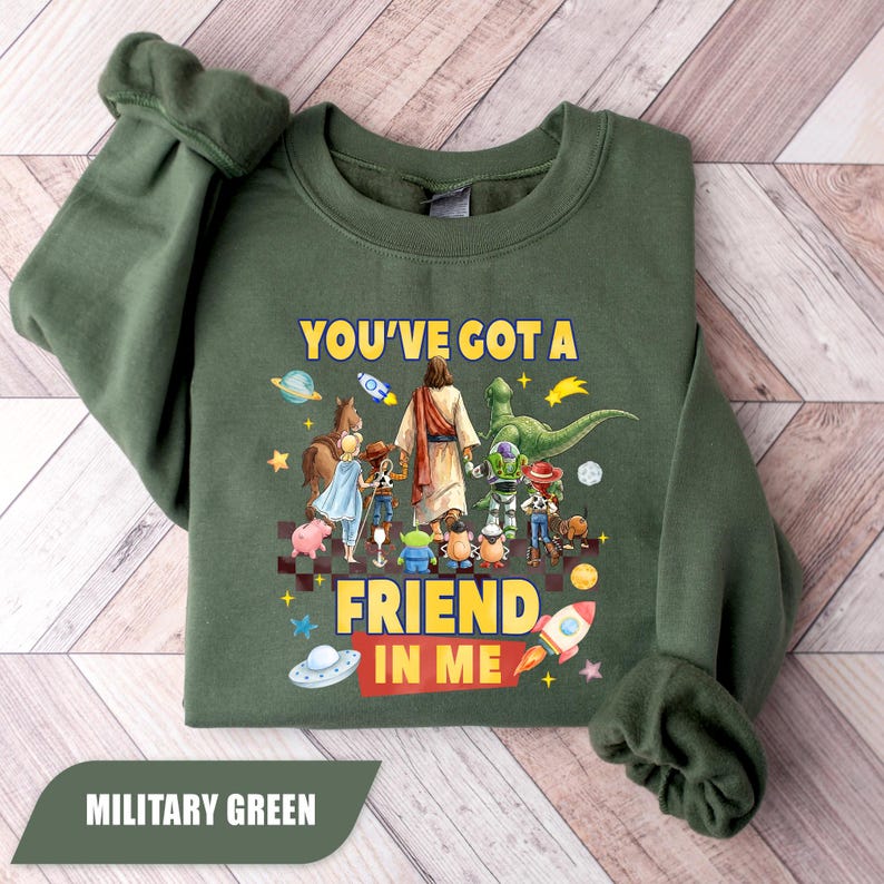 May include: A military green long-sleeve sweatshirt featuring a graphic print of a central figure in a robe surrounded by various animated toy characters. The text "YOU'VE GOT A FRIEND IN ME" is printed in yellow and red above and below the characters.