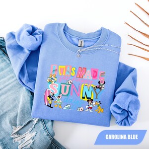 May include: Carolina blue sweatshirt with the text "I WAS MADE for SUNNY days" and cartoon characters. The sweatshirt has long sleeves and a crew neck. A silver chain necklace is on the sweatshirt. The sweatshirt is folded on top of a pair of blue jeans.