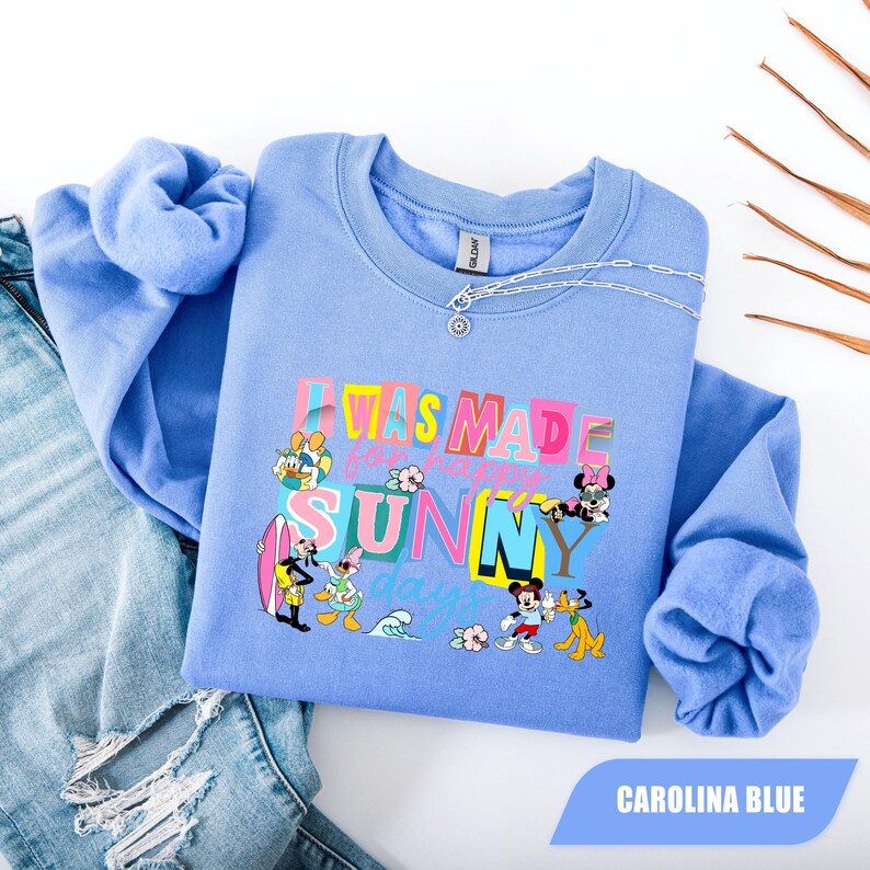 May include: Carolina blue sweatshirt with the text "I WAS MADE for SUNNY days" and cartoon characters. The sweatshirt has long sleeves and a crew neck. A silver chain necklace is on the sweatshirt. The sweatshirt is folded on top of a pair of blue jeans.