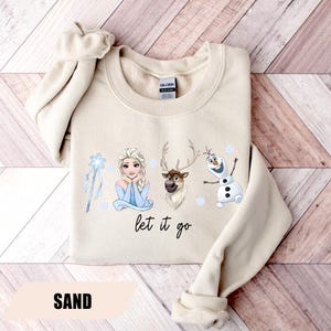 May include: Sand-colored sweatshirt featuring Elsa, a reindeer, and Olaf from Frozen, with the phrase "let it go." The design includes a snowflake wand and small blue snowflakes. The word "SAND" is printed at the bottom.
