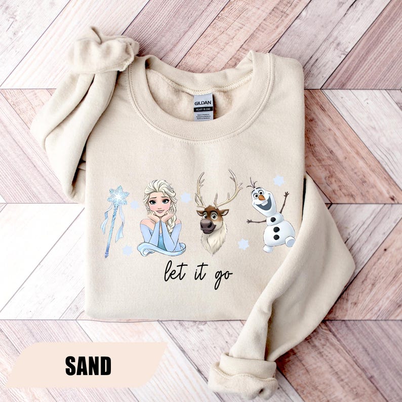 May include: Sand-colored sweatshirt featuring Elsa, a reindeer, and Olaf from Frozen, with the phrase "let it go." The design includes a snowflake wand and small blue snowflakes. The word "SAND" is printed at the bottom.