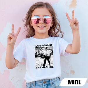 May include: White t-shirt with a black and white graphic and the text "RAGE AGAINST THE MACHINE". The shirt is worn by a person wearing pink sunglasses and making a peace sign with their fingers.