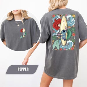 May include: A charcoal gray t-shirt featuring a small graphic on the front left chest and a large graphic on the back. Both prints show a blue alien character standing on a yellow surfboard next to a red-haired mermaid tail amidst ocean waves.
