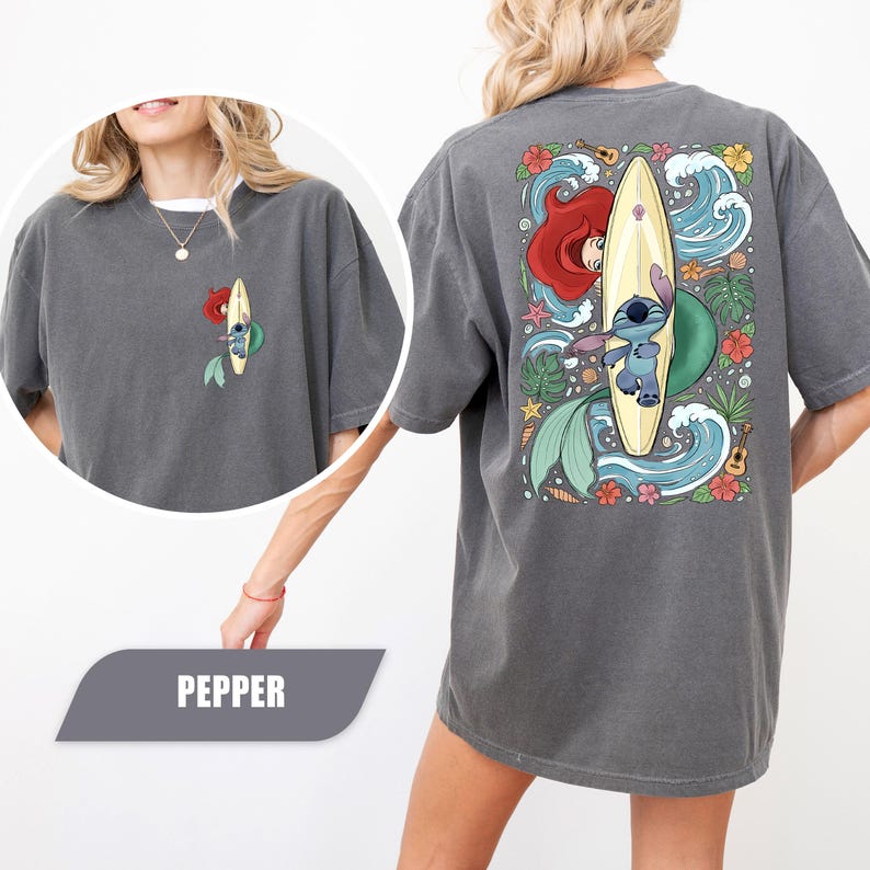 May include: A charcoal gray t-shirt featuring a small graphic on the front left chest and a large graphic on the back. Both prints show a blue alien character standing on a yellow surfboard next to a red-haired mermaid tail amidst ocean waves.