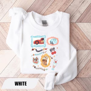 Puede incluir: Una sudadera blanca de cuello redondo con un estampado gr&aacute;fico de personajes de la pel&iacute;cula Cars, incluyendo a Rayo McQueen, Sally y Mate en marcos ornamentados, junto a ilustraciones de cerezas, un batido y cadenas de cuentas con texto.