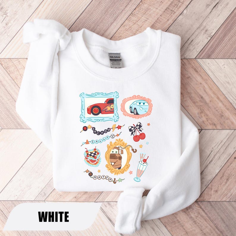 Puede incluir: Una sudadera blanca de cuello redondo con un estampado gr&aacute;fico de personajes de la pel&iacute;cula Cars, incluyendo a Rayo McQueen, Sally y Mate en marcos ornamentados, junto a ilustraciones de cerezas, un batido y cadenas de cuentas con texto.