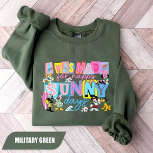 May include: Military green sweatshirt with the text "I WAS MADE for happy SUNNY days" in colorful block letters. Disney characters like Mickey Mouse, Donald Duck, and Minnie Mouse are featured in the design. The sweatshirt is a casual style.