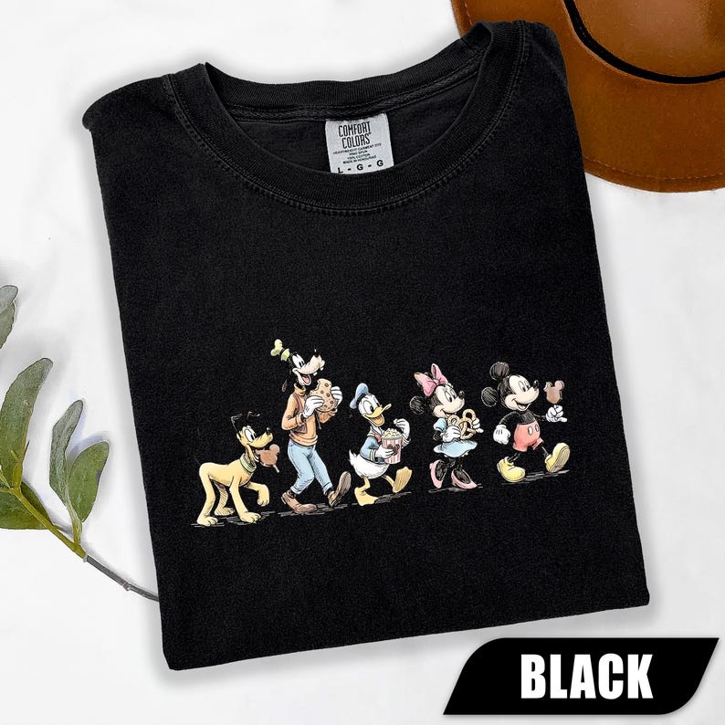 May include: A black t-shirt featuring a graphic print of five classic cartoon characters walking in a line. The characters include Pluto, Goofy, Donald Duck, Minnie Mouse, and Mickey Mouse, each holding a snack. The shirt has a Comfort Colors label.